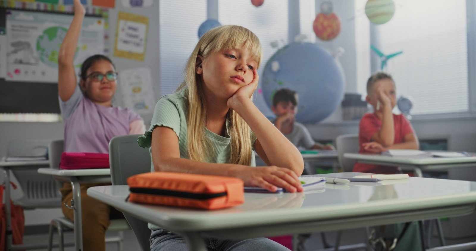 Young student sitting quietly at her classroom desk, looking discouraged while classmates participate in a lesson behind her.