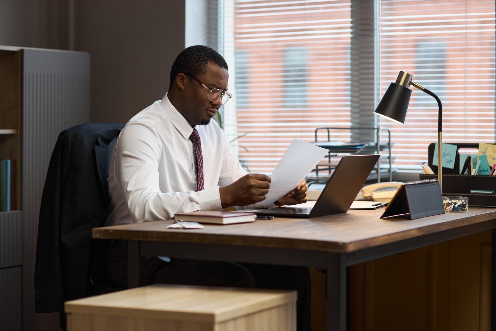 School administrator sitting at an office desk reviewing paperwork with a thoughtful expression.